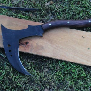 May include: A black curved axe with a wooden handle and white accents. The axe is lying on a wooden surface.