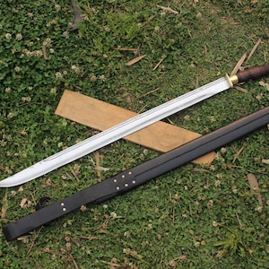 May include: A silver sword with a brown leather scabbard and a wooden handle. The sword is lying on a wooden surface with green grass in the background.