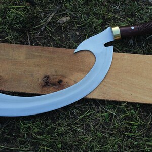 May include: A curved, silver-colored blade with a wooden handle and brass accents. The blade is shaped like a crescent moon and has a sharp point. The handle is wrapped in brown leather and has a brass pommel. The blade is resting on a piece of wood.