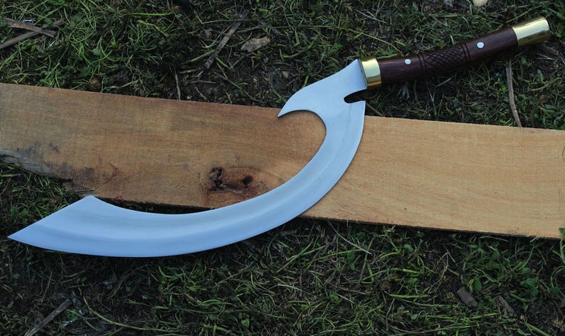 May include: A curved, silver-colored blade with a wooden handle and brass accents. The blade is shaped like a crescent moon and has a sharp point. The handle is wrapped in brown leather and has a brass pommel. The blade is resting on a piece of wood.