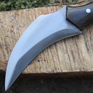 Puede incluir: Un cuchillo de hoja curva de color plateado con un mango marr&oacute;n oscuro. La hoja tiene un filo afilado y una forma curva. El mango es de madera oscura y tiene un peque&ntilde;o remache plateado. El cuchillo est&aacute; sobre una superficie de madera.