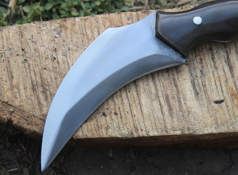 Puede incluir: Un cuchillo de hoja curva de color plateado con un mango marr&oacute;n oscuro. La hoja tiene un filo afilado y una forma curva. El mango es de madera oscura y tiene un peque&ntilde;o remache plateado. El cuchillo est&aacute; sobre una superficie de madera.