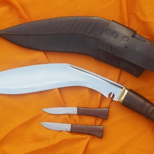 Könnte beinhalten: Ein großes, gebogenes Messer mit Holzgriff und zwei kleineren Messern, alle mit braunen Griffen und silbernen Klingen. Das Messer wird von einer schwarzen Lederscheide begleitet. Die Artikel werden auf einer orangefarbenen Oberfläche präsentiert.