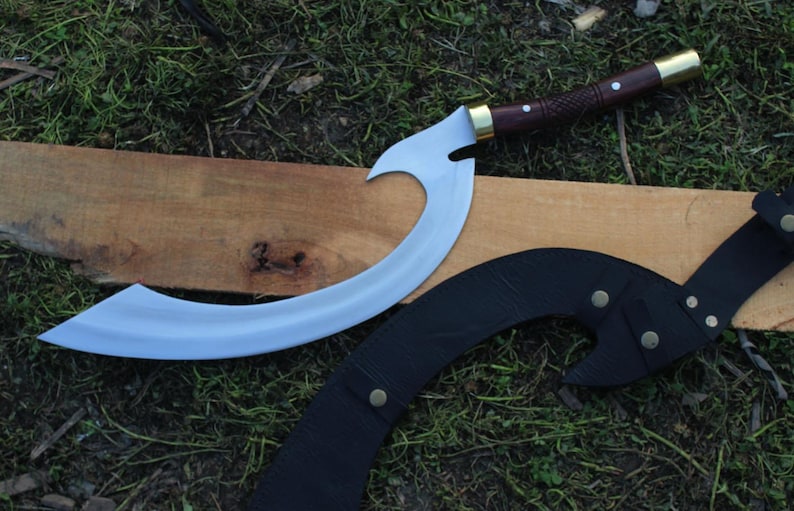 May include: A curved, silver-colored knife with a wooden handle and brass accents. The knife is resting on a wooden surface with a black leather sheath.