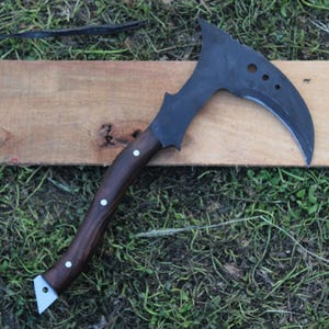 May include: A black and brown curved axe with a wooden handle. The axe is resting on a wooden surface.