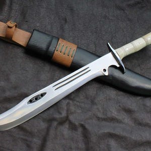 May include: A silver-colored blade with a curved edge and a light-colored handle rests on a black sheath. The sheath is accented with brown leather and stitching. The knife has three parallel grooves along the blade.