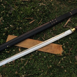 Pu&ograve; includere: Una lunga spada argentata con un fodero in pelle nera. La spada ha un'impugnatura in legno con un pomo in ottone e una guardia in ottone. La spada &egrave; appoggiata su una superficie in legno marrone.