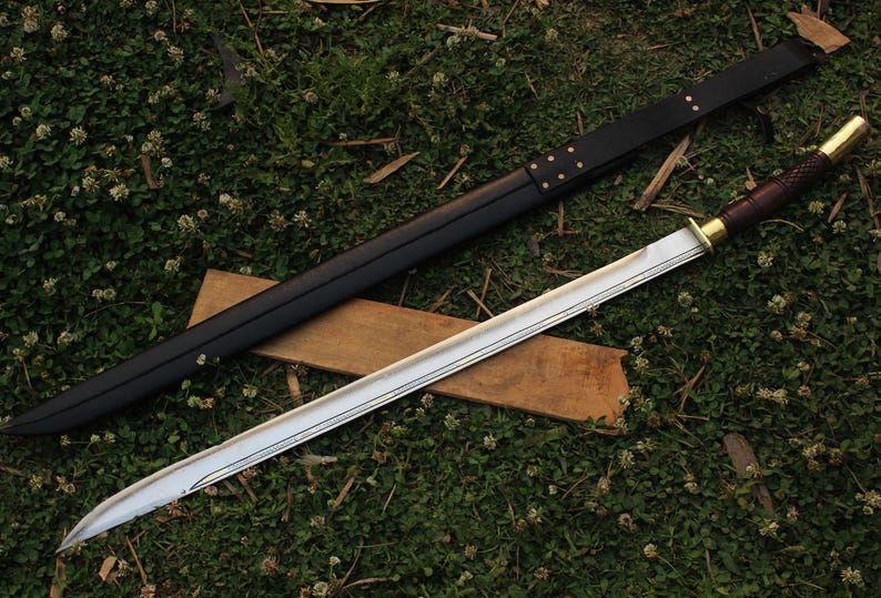 Pu&ograve; includere: Una lunga spada argentata con un fodero in pelle nera. La spada ha un'impugnatura in legno con un pomo in ottone e una guardia in ottone. La spada &egrave; appoggiata su una superficie in legno marrone.