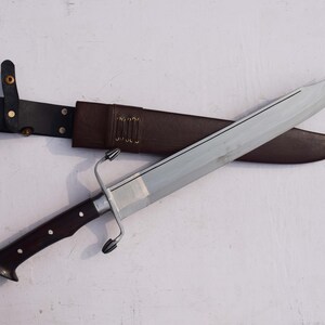 May include: A large, silver-bladed knife with a dark wooden handle and a brown leather sheath. The knife features a crossguard and is designed for combat or display. The sheath has a belt loop for carrying.