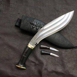 May include: A large, curved blade knife with a silver finish and a black handle, resting on a dark surface. The knife is accompanied by a black sheath and two smaller knives. The blade has a distinctive curved shape and a decorative design.