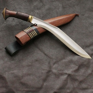 May include: A traditional kukri knife with a curved blade, resting on its wooden sheath. The handle is dark brown with brass accents. The sheath is also brown with leather and brass detailing. The knife and sheath are set against a dark background.