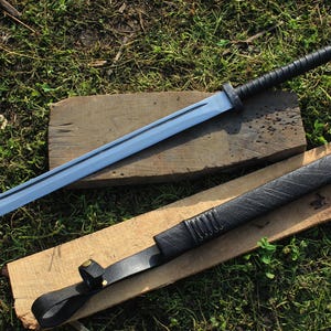 May include: A long, silver-colored sword with a black leather handle and a black leather scabbard. The sword is lying on a wooden surface.