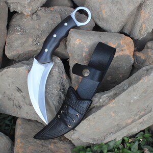 Puede incluir: Un cuchillo karambit curvo con mango negro y hoja plateada, junto a una funda de cuero negro. El cuchillo tiene un anillo para el dedo al final del mango. La funda tiene un patr&oacute;n decorativo y una correa con cierre de bot&oacute;n.