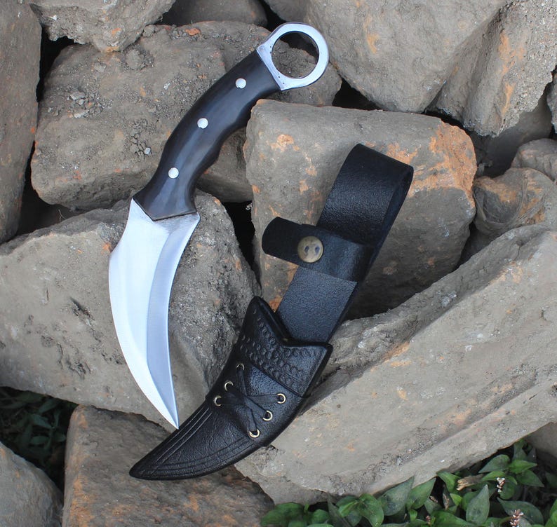 Puede incluir: Un cuchillo karambit curvo con mango negro y hoja plateada, junto a una funda de cuero negro. El cuchillo tiene un anillo para el dedo al final del mango. La funda tiene un patr&oacute;n decorativo y una correa con cierre de bot&oacute;n.