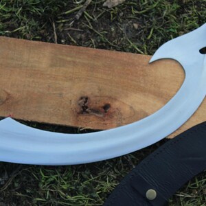 May include: A curved, silver-colored blade with a wooden handle and brass accents. The blade is resting on a wooden surface and a black leather sheath.