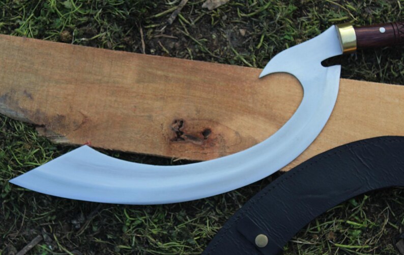 May include: A curved, silver-colored blade with a wooden handle and brass accents. The blade is resting on a wooden surface and a black leather sheath.