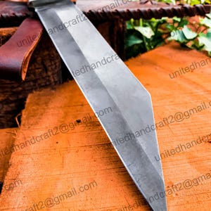 Op de afbeelding: Een close-up van een zilveren mesblad met een scherpe punt. Het blad rust op een houten oppervlak. Het blad heeft een lichte kromming en is erg scherp.