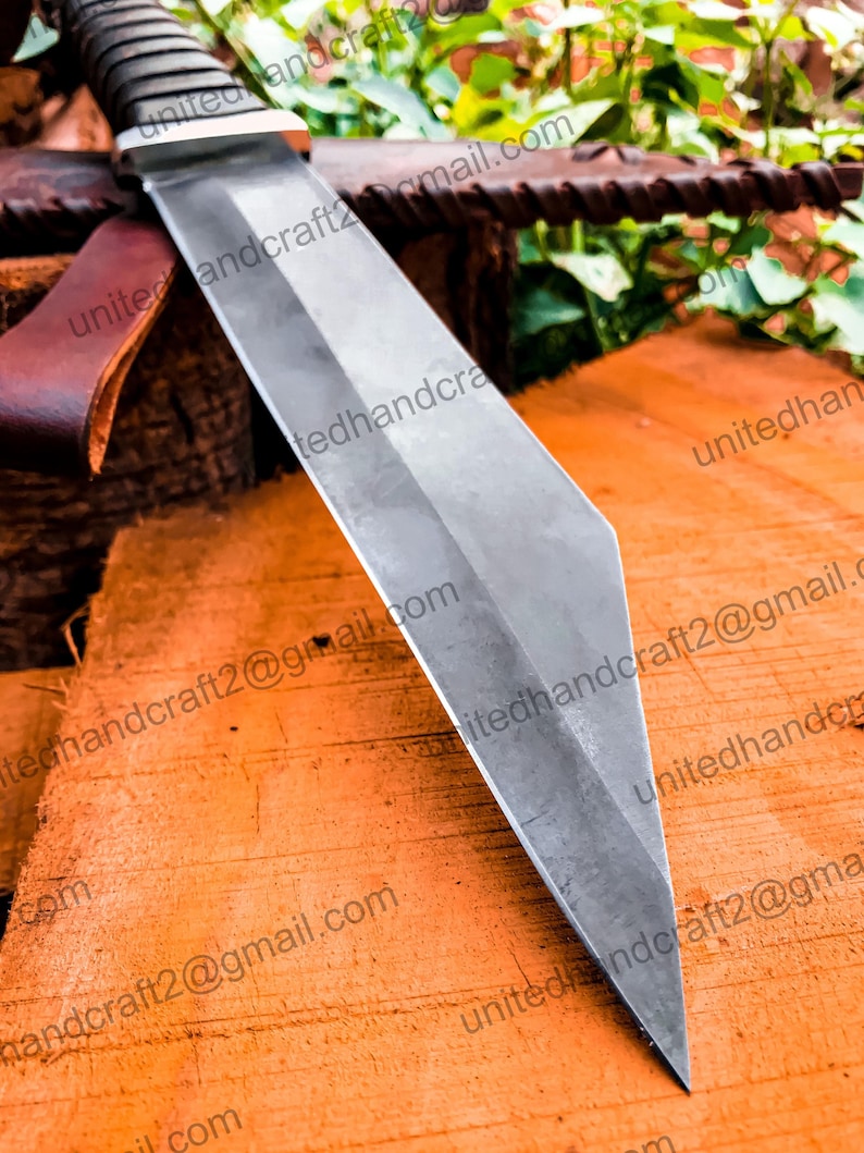 Op de afbeelding: Een close-up van een zilveren mesblad met een scherpe punt. Het blad rust op een houten oppervlak. Het blad heeft een lichte kromming en is erg scherp.
