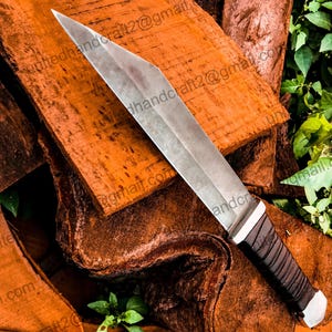 Op de afbeelding: Een zilveren mes met een zwart leren handvat, liggend op een stapel bruin hout. Het mes heeft een scherpe punt en een recht blad.