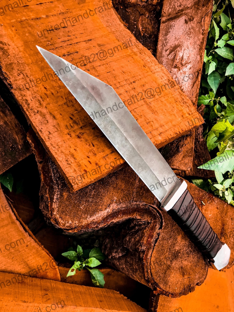 Op de afbeelding: Een zilveren mes met een zwart leren handvat, liggend op een stapel bruin hout. Het mes heeft een scherpe punt en een recht blad.