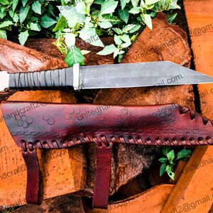 Op de afbeelding: Een mes met een zwarte handgreep en een leren schede. De schede heeft een decoratief patroon en wordt vastgezet met een band. Het mes ligt op een stapel hout.