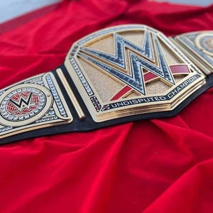 May include: A gold and black championship belt with the letters "W" and "WWE" on the front. The belt has red and silver accents and the words "Undisputed Champion" are printed on the front. The belt is lying on a red surface.