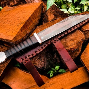 Op de afbeelding: Een zilveren en zwart mes met een leren schede. Het mes ligt op een stapel hout.
