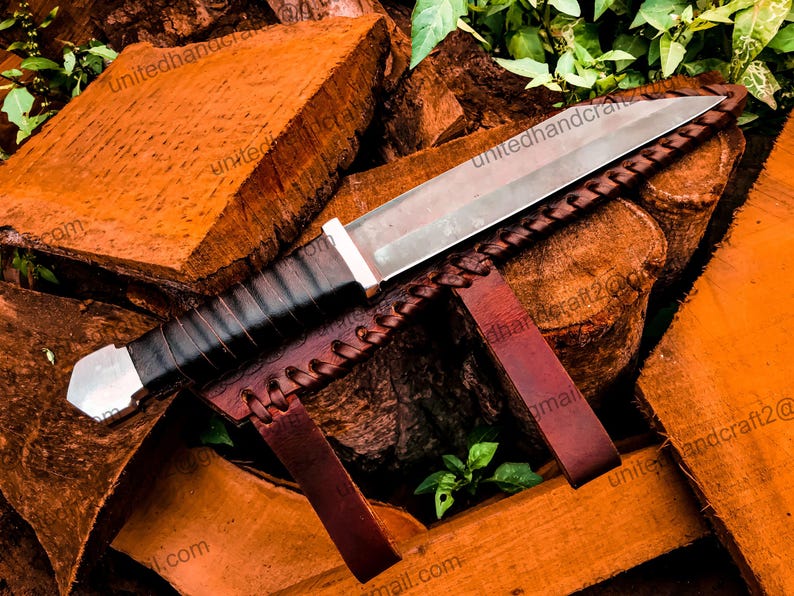 Op de afbeelding: Een zilveren en zwart mes met een leren schede. Het mes ligt op een stapel hout.