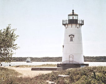 May include: A white lighthouse with a black roof and a black railing stands on a sandy beach with a path leading to it. The lighthouse is surrounded by green bushes and a body of water is visible in the background.