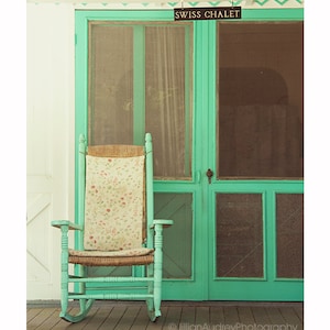 May include: A rocking chair with a floral patterned cushion sits on a wooden porch in front of a turquoise green door with a sign that reads "Swiss Chalet".
