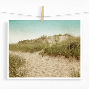 May include: A print of a sandy path leading through a dune covered in green grass, under a light blue sky. The print is held by a wooden clothespin on a white string, creating a rustic, beachy aesthetic.