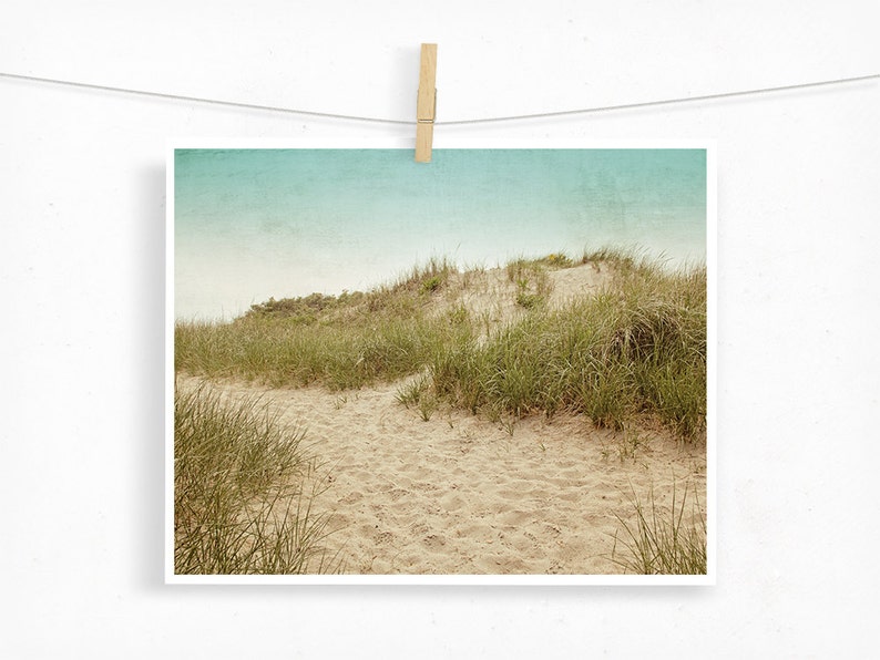 May include: A print of a sandy path leading through a dune covered in green grass, under a light blue sky. The print is held by a wooden clothespin on a white string, creating a rustic, beachy aesthetic.