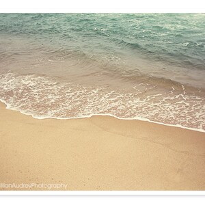 May include: A close-up of a sandy beach with foamy waves lapping at the shore. The water is a light blue-green color, and the sand is a warm beige.
