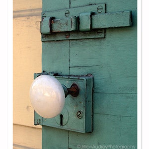 May include: A white porcelain door knob with a brass latch on a teal green wooden door. The door has a metal latch with a rusty finish.