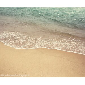 May include: A close-up of a sandy beach with a foamy wave lapping at the shore. The water is a light blue-green color, and the sand is a warm beige.