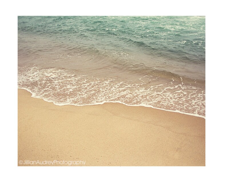 May include: A close-up of a sandy beach with a foamy wave lapping at the shore. The water is a light blue-green color, and the sand is a warm beige.