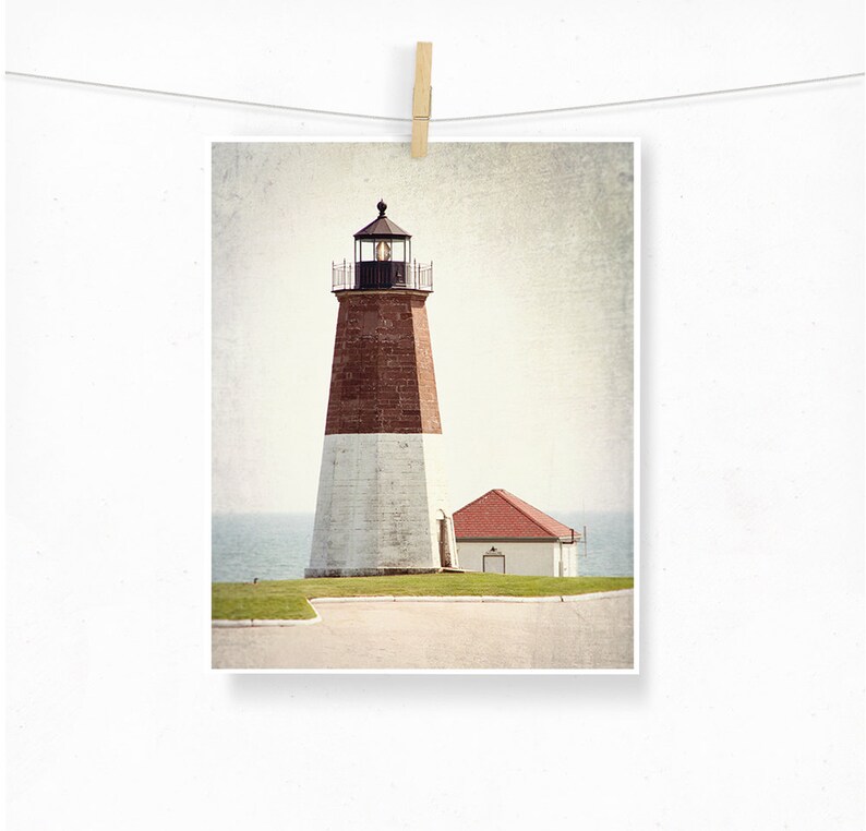 May include: A vintage-style photograph of a red and white lighthouse with a smaller building next to it. The lighthouse is on a grassy hill with a paved road leading to it. The sky is a light blue with some clouds.