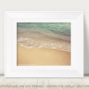 May include: A photograph of a beach scene with blue water and white waves crashing on the shore. The sand is a light tan color.