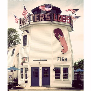May include: A white building with a red lobster sign and the words "Lobsters Lobsters" on the side. The building has a red lobster painted on the side and the word "Fish" underneath. There are American flags hanging from the building.