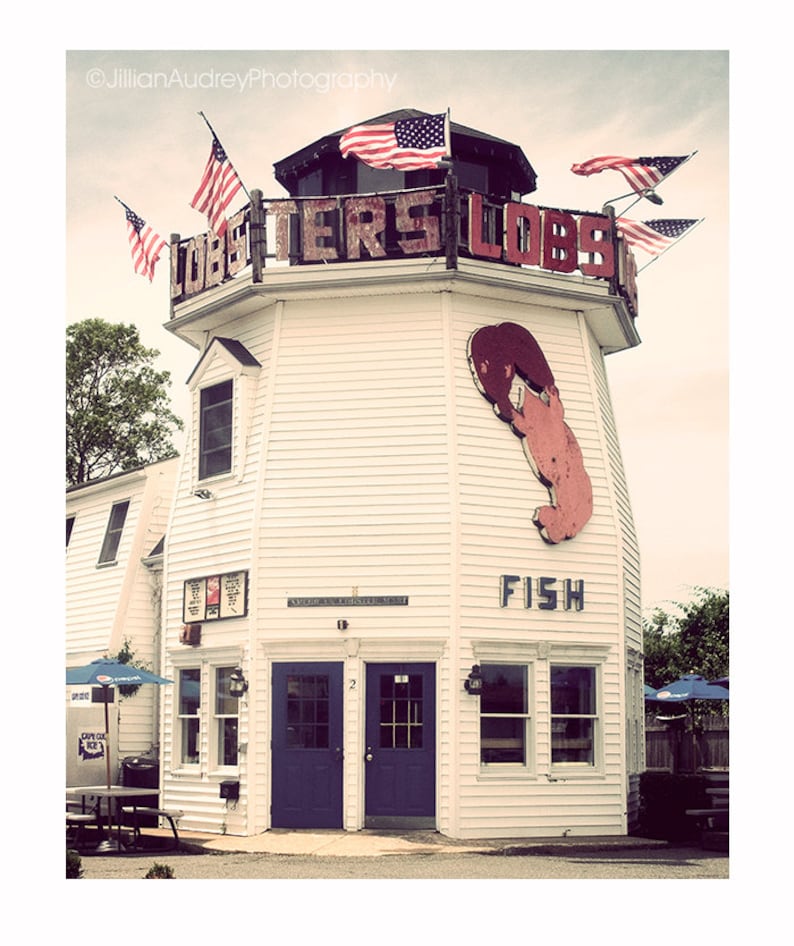 May include: A white building with a red lobster sign and the words "Lobsters Lobsters" on the side. The building has a red lobster painted on the side and the word "Fish" underneath. There are American flags hanging from the building.