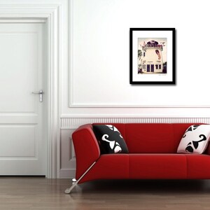 May include: A framed photograph of a white building with a red roof and a blue door. The building has a balcony and a sign that says "The Pier House". The photograph is hanging on a white wall above a red couch.