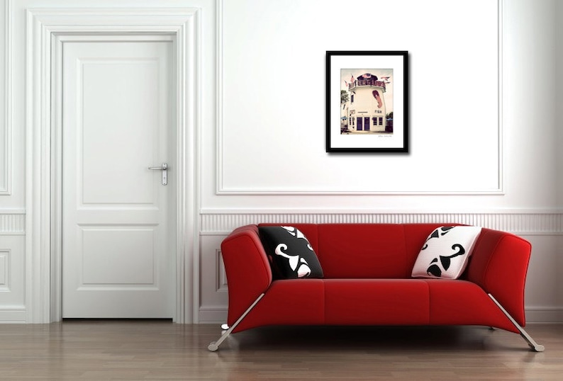 May include: A framed photograph of a white building with a red roof and a blue door. The building has a balcony and a sign that says "The Pier House". The photograph is hanging on a white wall above a red couch.