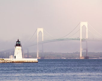 May include: A white lighthouse with a black roof stands on a small island in front of a large suspension bridge. The bridge is white with green supports and a green roadway. The scene is hazy and the water is a light blue.