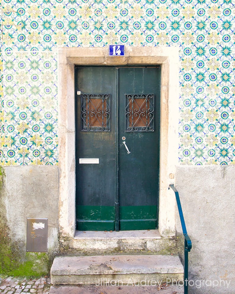 May include: A green wooden door with a number "14" above it. The door has two windows with black metal bars and a silver door handle. The door is set in a white frame and is surrounded by a wall covered in blue and white patterned tiles.