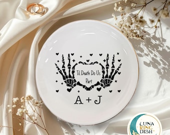 May include: White ceramic ring dish with a gold rim, featuring black skeleton hands forming a heart around the text "Til Death Do Us Part" and initials "A + J". Decorative hearts are scattered around the design. Two gold rings are visible.