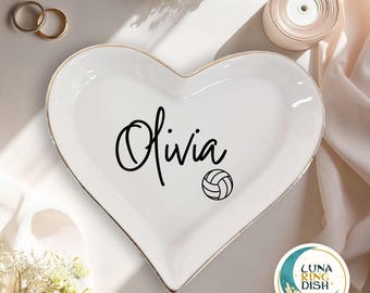 May include: White heart-shaped ceramic ring dish with gold trim. The dish features the name "Olivia" in black script, along with a volleyball graphic. Two gold rings and a roll of ribbon are visible in the background.