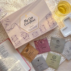 May include: A pink box with the words "My Cup Overflows" and a design of teacups with bible verses. The box contains individual tea bags with bible verses printed on them. The tea bags are arranged on a white surface with a white cross and a glass of tea.