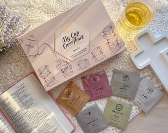 May include: A pink box with the words "My Cup Overflows" and a design of teacups with bible verses. The box contains individual tea bags with bible verses printed on them. The tea bags are arranged on a white surface with a white cross and a glass of tea.