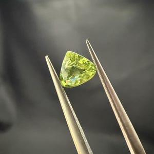 Pode incluir: Uma gema verde com forma triangular &eacute; segurada por pin&ccedil;as. A gema tem um leve tom amarelo e &eacute; transparente.