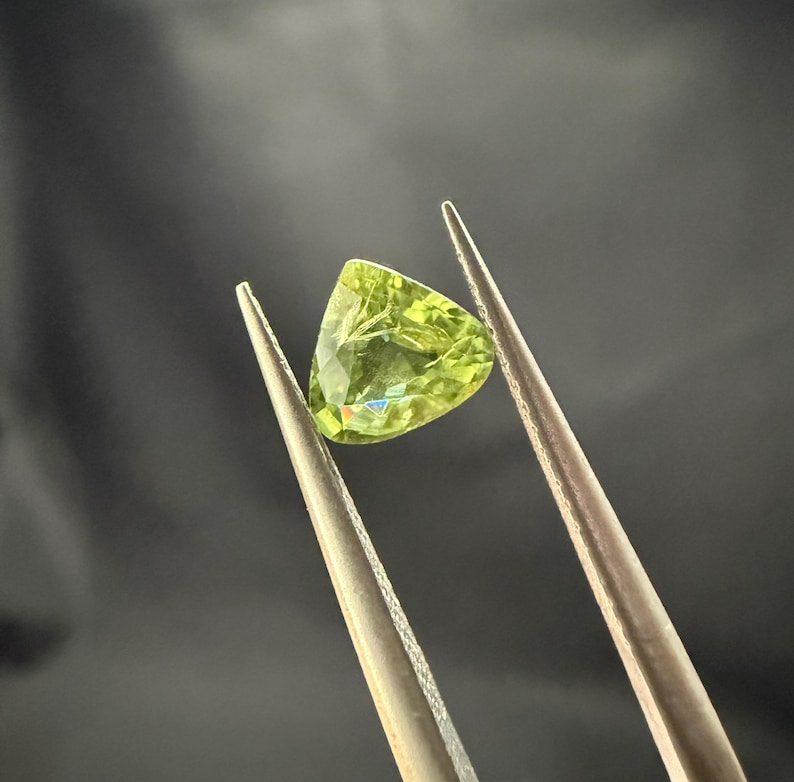 Pode incluir: Uma gema verde com forma triangular &eacute; segurada por pin&ccedil;as. A gema tem um leve tom amarelo e &eacute; transparente.